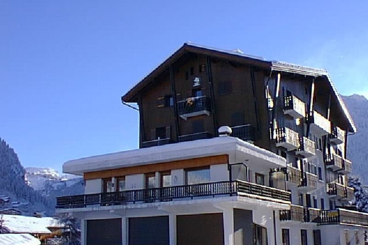A snow-covered hotel with a modern white base and a traditional brown chalet-style upper section, set against a clear blue sky and snowy mountains.