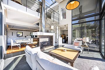 Bright, modern open-plan living space with high ceilings, glass walls, a stone fireplace, and a mezzanine. Includes dining and kitchen areas.