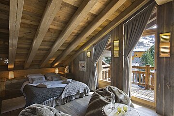 A cozy, rustic wooden attic bedroom with exposed beams, a plush bed, and soft furnishings. A balcony door reveals a view of a village and trees.