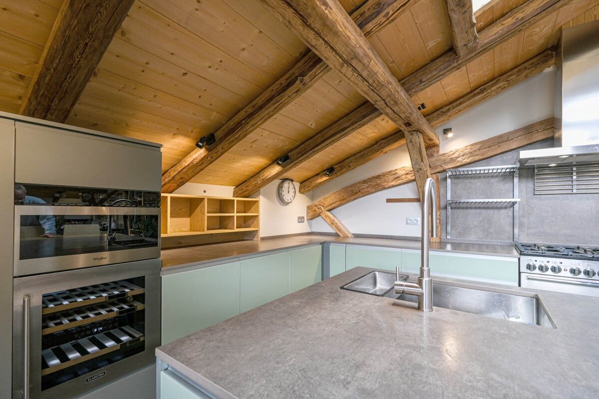 A modern kitchen with a sloped wooden ceiling and exposed beams. Features light green cabinets, gray countertops, a sink, stove, oven, and wine fridge.
