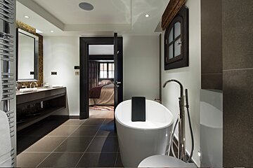 Modern luxury bathroom featuring a freestanding white bathtub, vanity with large mirror, and an open doorway leading to a bedroom.