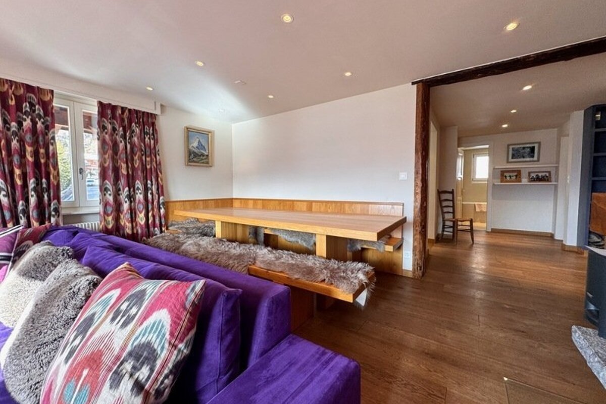 A cozy chalet living room featuring a bright purple sofa, patterned pillows, and a wooden dining table with fur-lined benches.