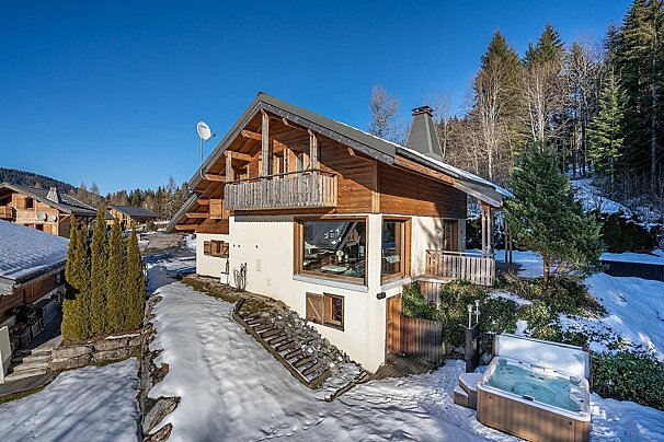 A rustic wooden and white chalet sits in a snowy mountain landscape under a clear blue sky, with a hot tub on the patio.