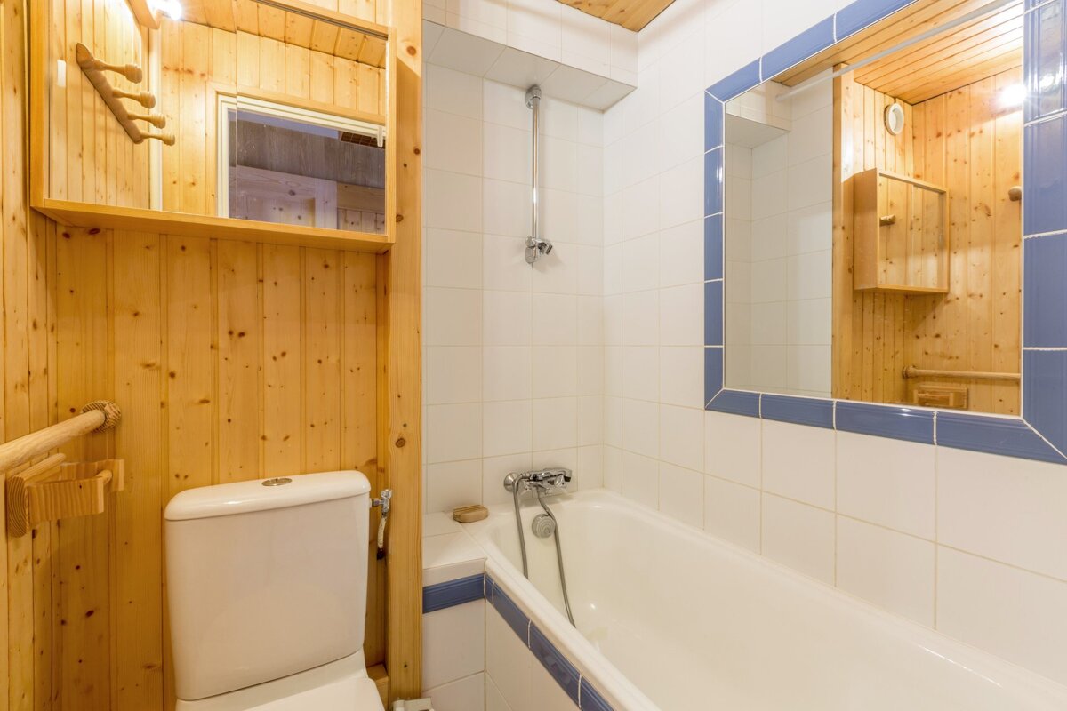A compact bathroom with a rustic feel, featuring wood paneling on one wall and white tiles with blue trim on the other, a toilet, a bathtub with a shower, and a mirror.