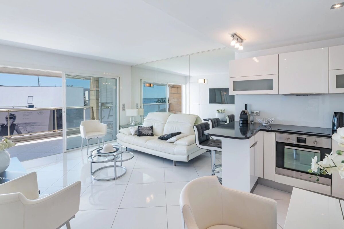A bright, modern white open-plan living room and kitchen with glossy floors, sleek furniture, and a balcony with a scenic view.