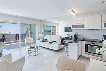 Bright, modern apartment interior with a white leather sofa, open-plan kitchen, and polished tiled flooring leading to a balcony.