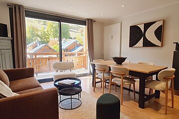 Cozy living and dining area with modern wooden furniture, a brown sofa, and large windows looking out onto scenic wooden cabins.