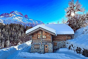 A picturesque snowy wooden chalet with a colorful stone base, surrounded by snow-covered mountains, a pine forest, and a brilliant blue sky.
