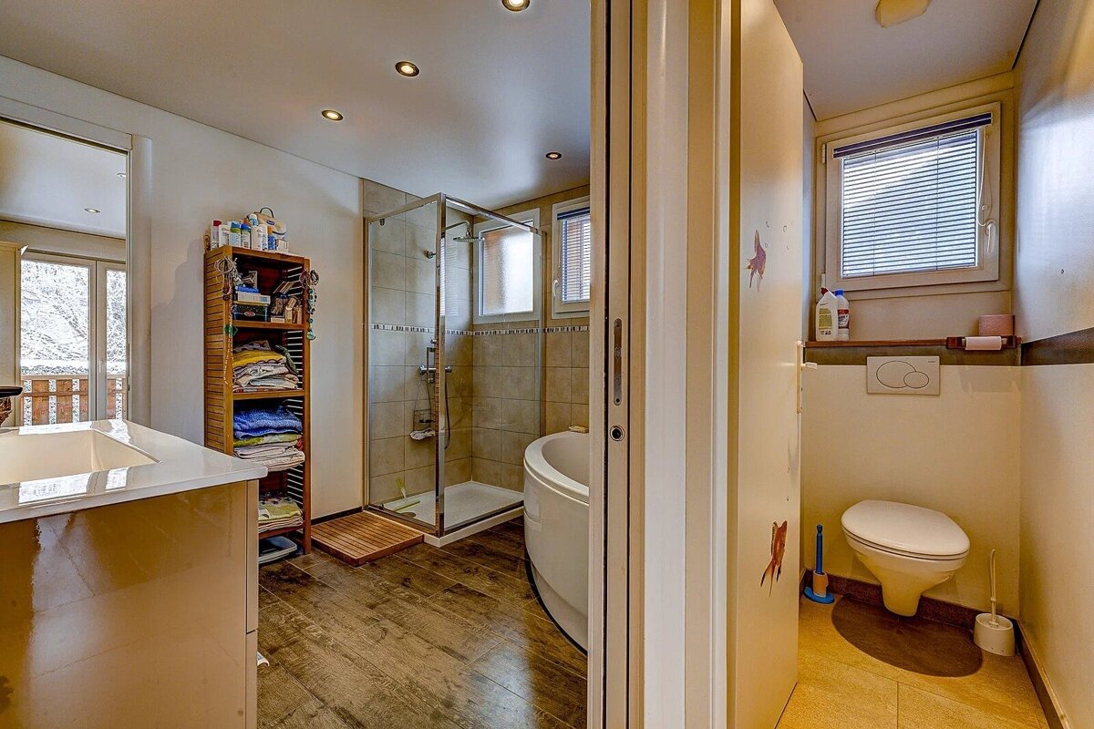 A cozy bathroom with a shower, round tub, vanity, and toilet alcove. It features warm wooden flooring and a window looking out to a snowy view.