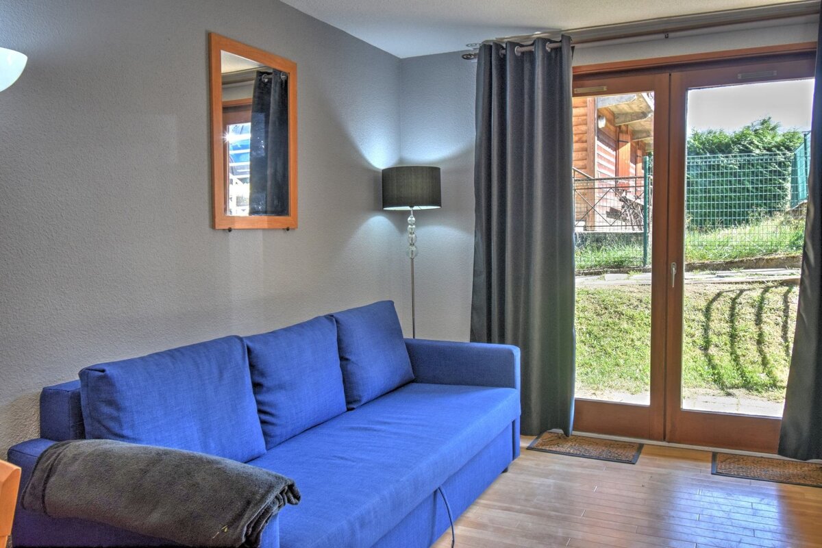 A cozy living area with a bright blue sofa, grey walls, a floor lamp, and a mirror. A glass door opens to a green lawn outside.