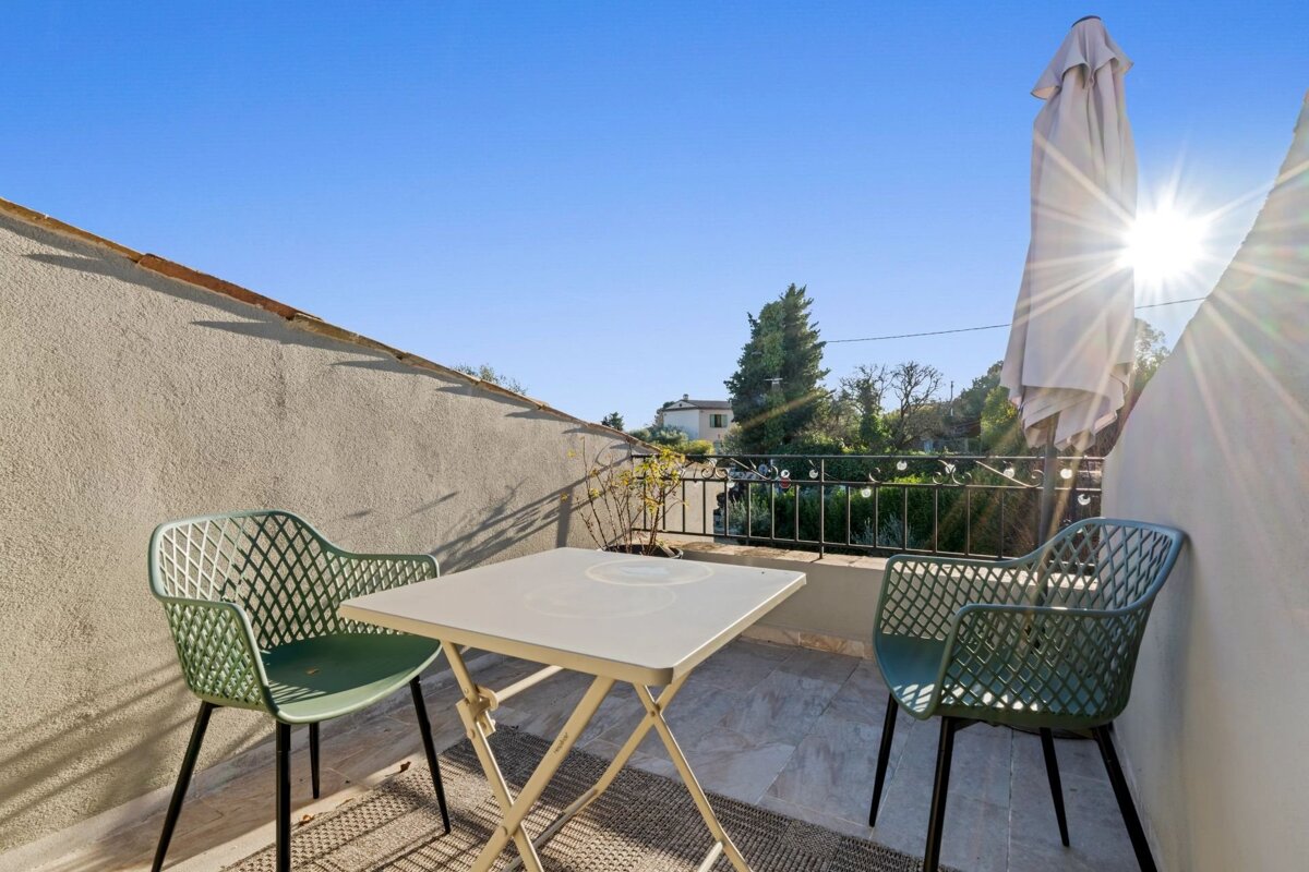 Sunny balcony with a small white table, two green mesh chairs, a closed patio umbrella, and bright sun shining in a blue sky.