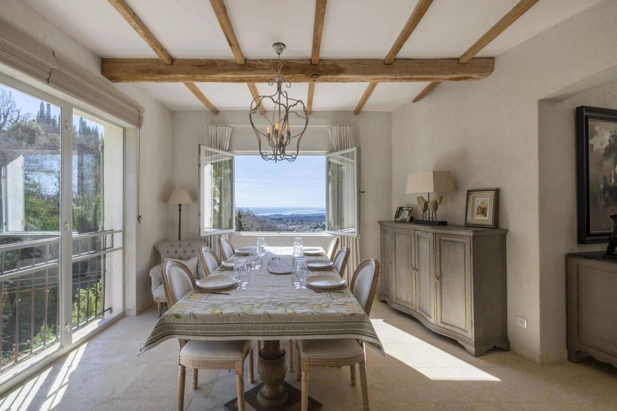 A charming dining room featuring a set table, wooden beams, an ornate chandelier, and large windows with a breathtaking panoramic view.
