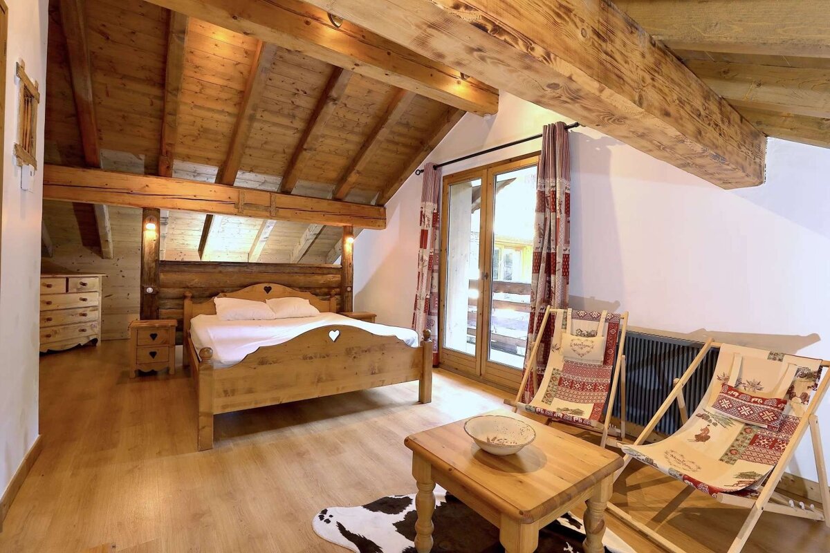 A rustic wooden-beamed attic bedroom with a heart-carved bed, balcony, two patterned deck chairs, and a small table on a cowhide rug.