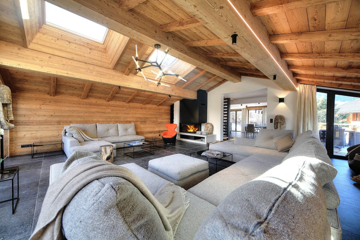 A spacious, modern chalet-style living room with a vaulted wood ceiling, skylights, large grey sofas, a cozy fireplace, and an orange accent chair.