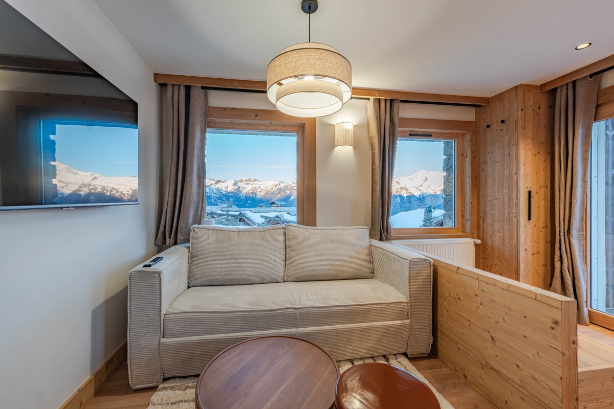 Cozy chalet living room with a beige sofa, wooden details, and windows framing a beautiful view of snowy mountains.