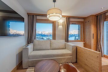 Cozy chalet living room with a beige sofa, wooden details, and windows framing a beautiful view of snowy mountains.