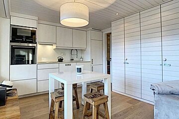 Minimalist white kitchen featuring a square dining table with wooden stools, modern cabinetry, and a large pendant light.