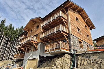 A multi-story wooden chalet with balconies, built on a stone foundation next to a pine forest under a cloudy sky.