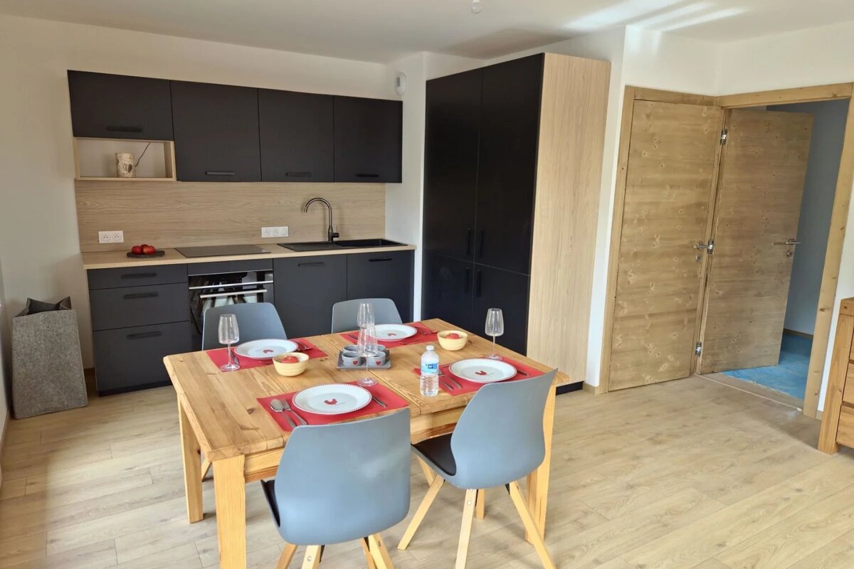 Modern kitchen and dining area featuring a wooden table set for four with grey chairs, black cabinets, and light wood finishes.