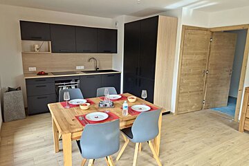 Modern kitchen and dining area featuring a wooden table set for four with grey chairs, black cabinets, and light wood finishes.