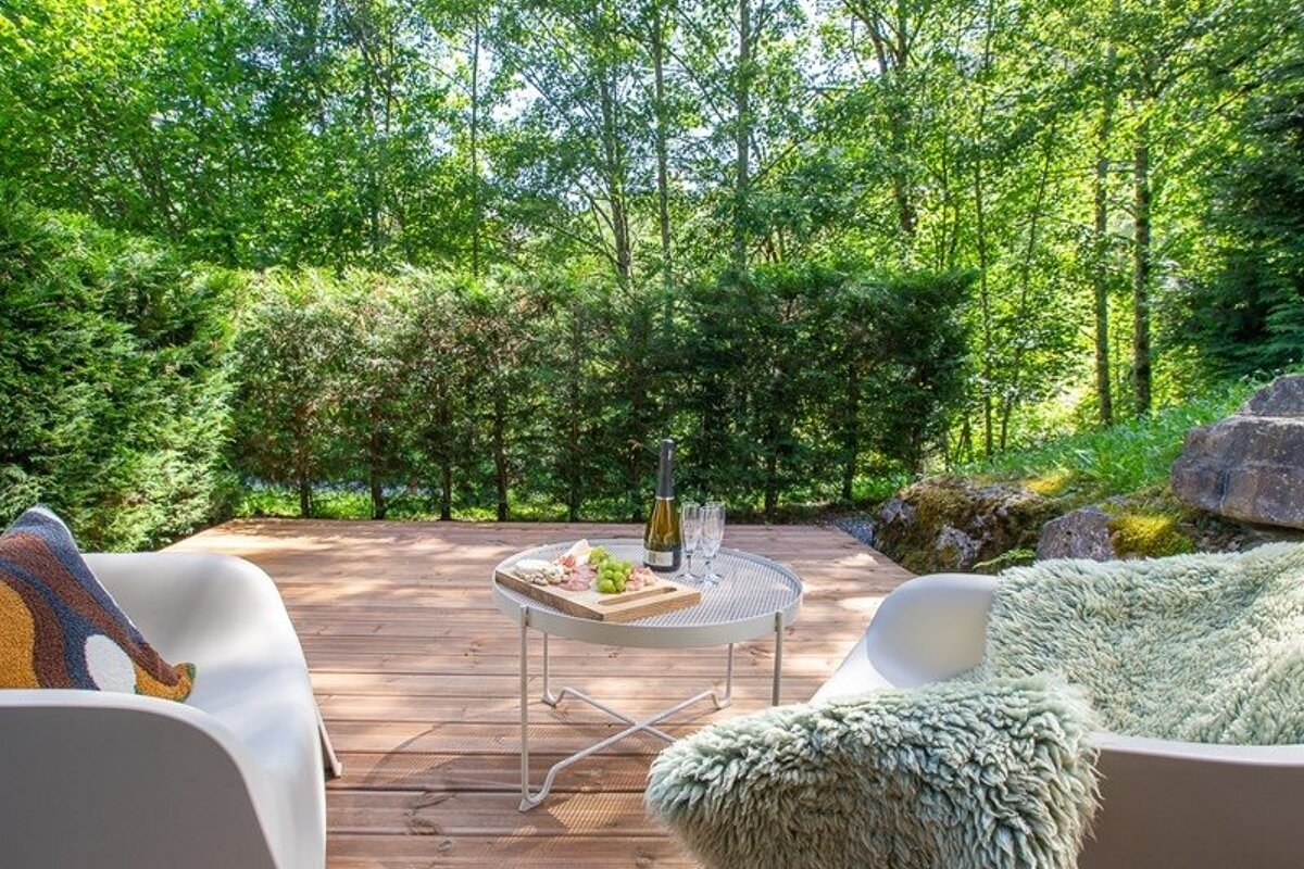 A cozy wooden deck in a lush forest with lounge chairs and a small table set with wine, glasses, and a cheese board.