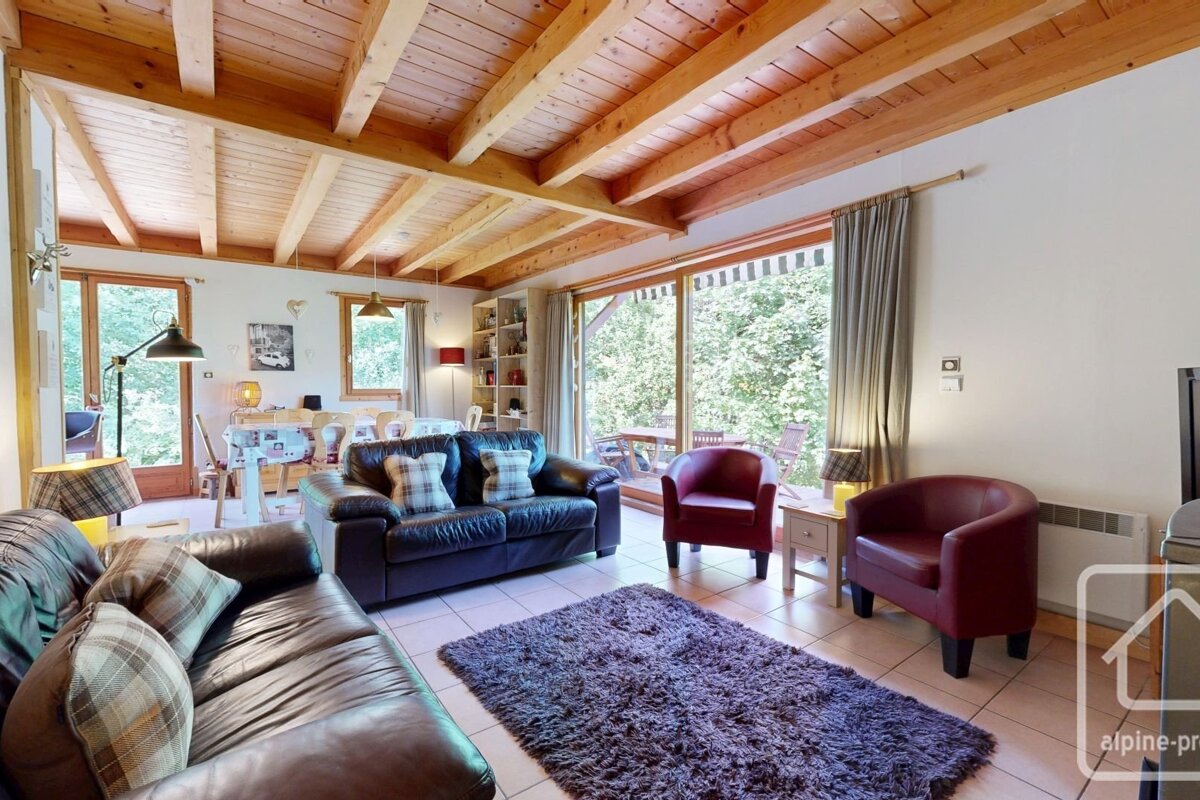 Cozy chalet living room with wooden beamed ceiling, leather sofas, red armchairs, and a shaggy rug, overlooking a lush green outdoor area.