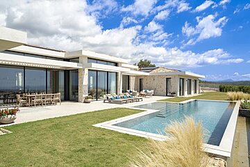 Modern luxury villa featuring a swimming pool, green lawn, and outdoor seating under a bright, cloudy sky.