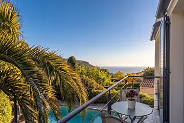 A sunny balcony with a table, overlooking a swimming pool, lush greenery, and the sea in the distance.