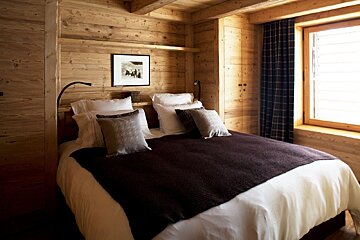A rustic, wood-paneled bedroom features a cozy bed with white and brown bedding, framed art, and a window with a plaid curtain.