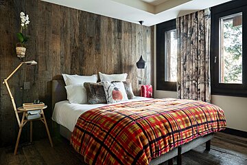 Cozy, rustic bedroom with a wood-paneled wall, red plaid duvet on the bed, and large windows revealing a snowy outdoor scene.