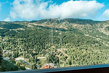 A panoramic view of a vast, forested mountain landscape under a cloudy blue sky, with a village nestled among the trees in the valley.