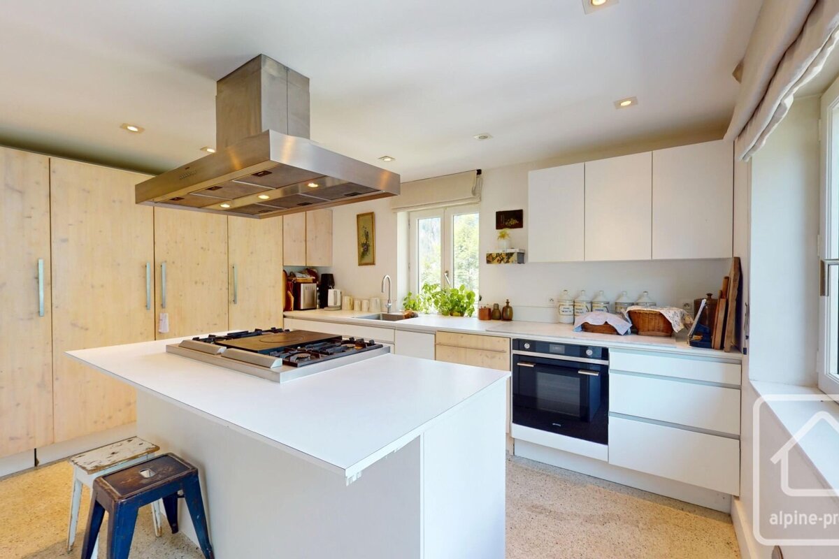 Bright modern kitchen with pale wood cabinets, a white island with cooktop, stainless steel hood, and built-in oven.
