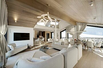 Modern chalet interior with light wood ceilings, plush white sofas, a stylish chandelier, and large windows showcasing snowy mountain views.
