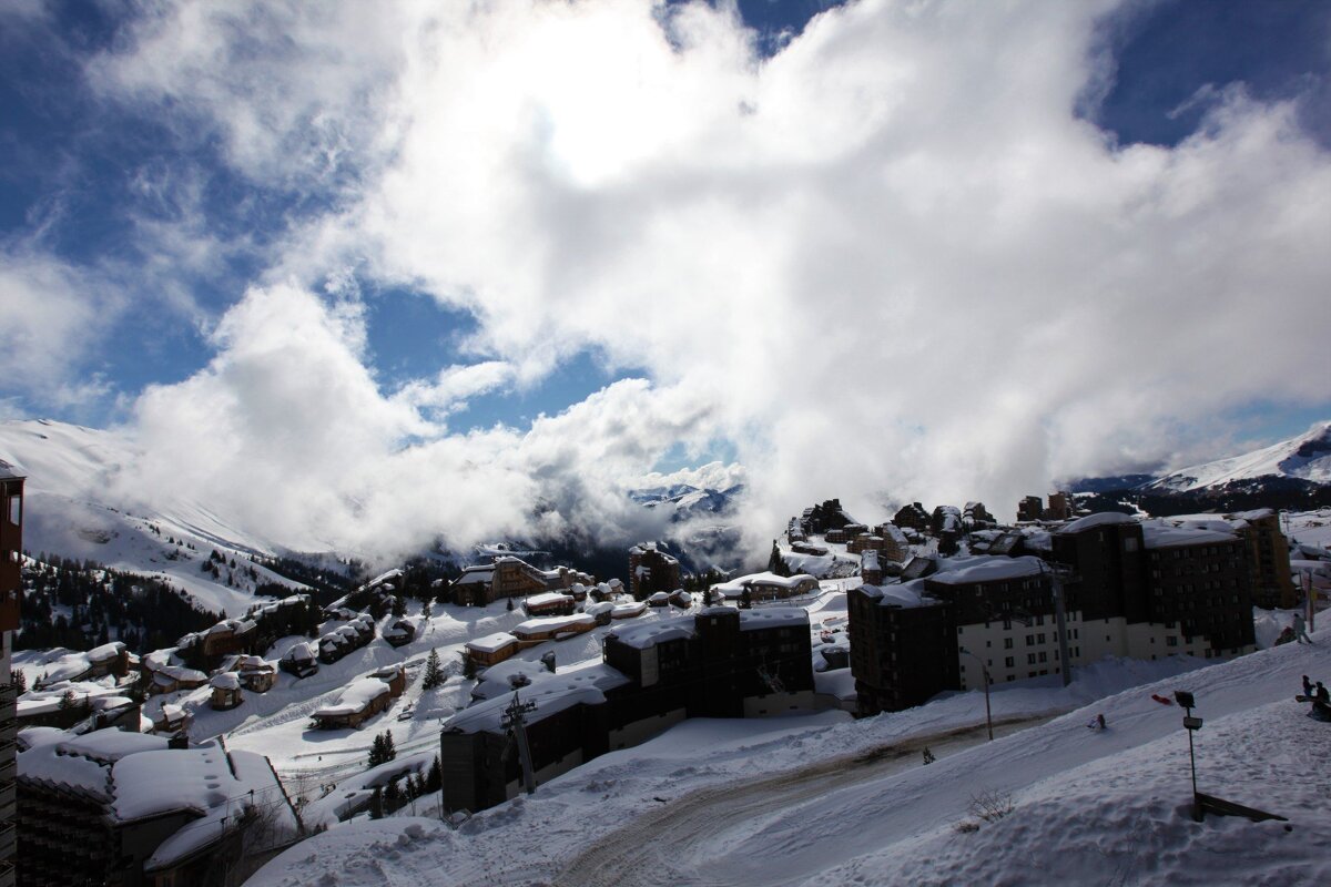 A snow-covered ski resort village with numerous buildings nestled on snowy mountain slopes, partially shrouded by dramatic clouds against a blue sky.