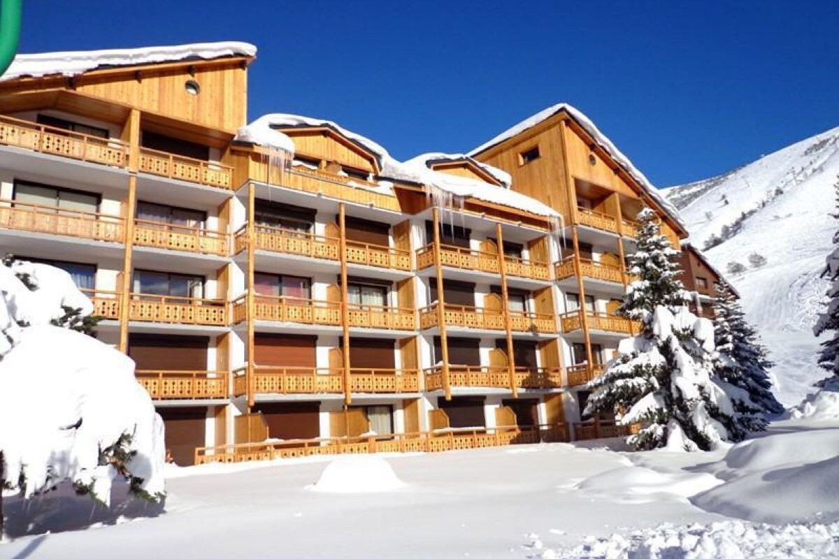 A large ski lodge with wooden balconies is covered in snow under a clear blue sky. Icicles hang from the roof, surrounded by snowy trees and mountains.