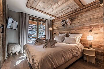 Cozy ski chalet bedroom with warm wooden walls/ceiling, a plush bed, a TV, and a scenic snowy mountain village view outside the window.