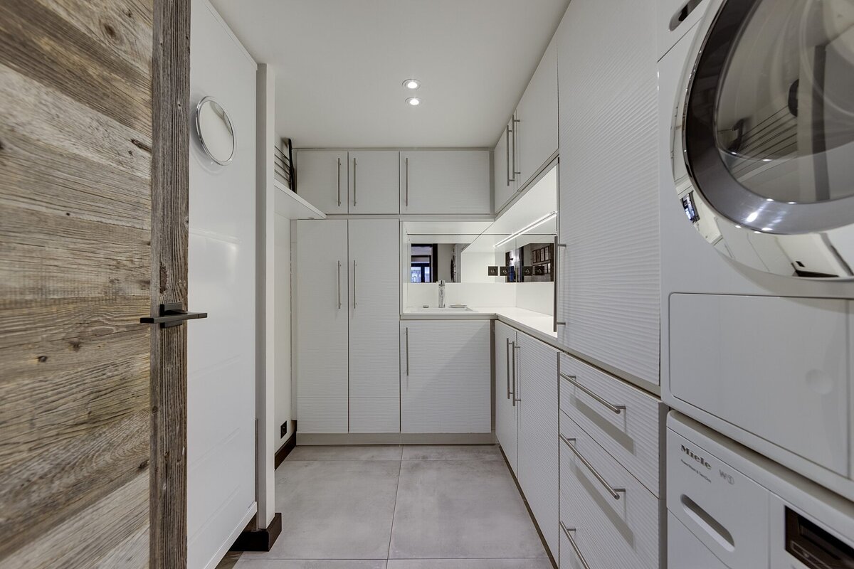 A sleek, modern laundry room featuring white cabinetry, a washer/dryer, and a compact sink area. A rustic wooden door frames the left.