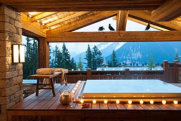 Candlelit hot tub on a wooden deck overlooking snowy mountains, with champagne, a bench, and birds perched on overhead timber beams.