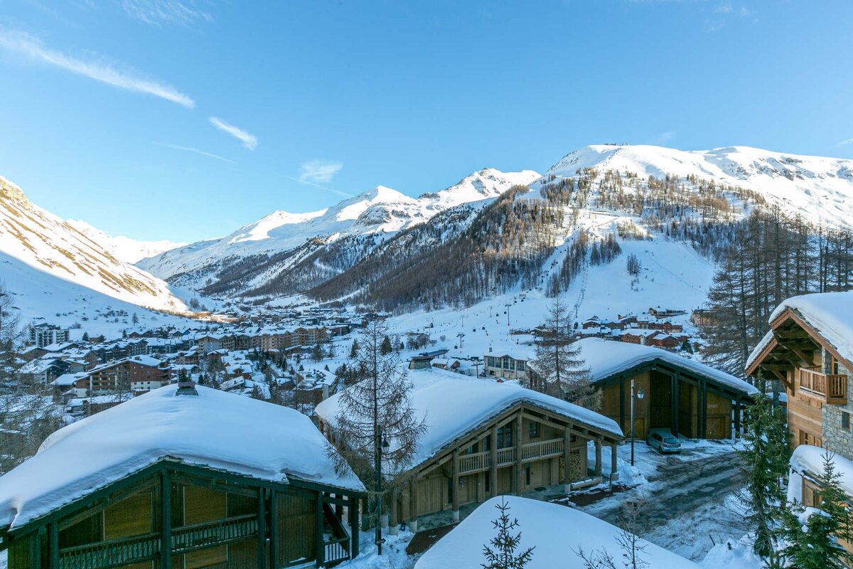 A scenic snowy alpine village with wooden chalets, surrounded by majestic snow-covered mountains and ski slopes under a bright blue sky.