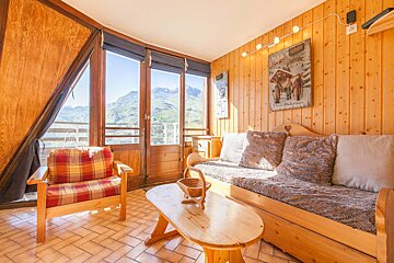 A rustic, sunlit, wood-paneled room with wooden furniture and large windows framing a stunning mountain view.