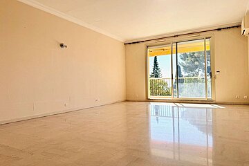 Bright, empty room with light walls, glossy floor, and a large sliding glass door revealing a balcony with trees and abundant sunlight.