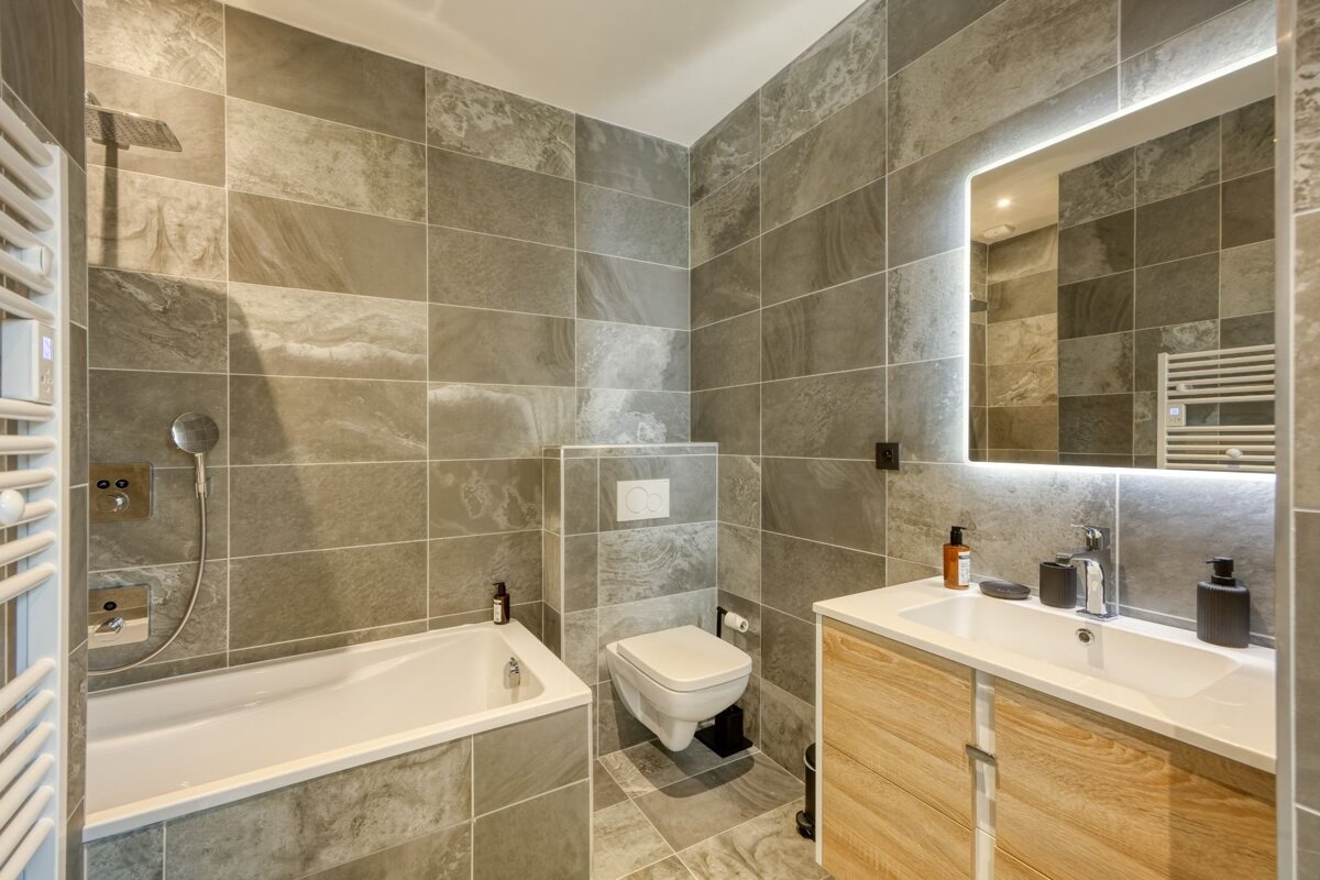 A sleek, modern bathroom with grey tiled walls and floor, featuring a bathtub, toilet, wood vanity with sink, and a backlit mirror.
