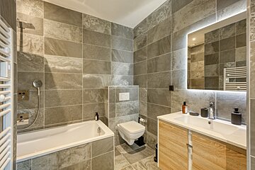 A sleek, modern bathroom with grey tiled walls and floor, featuring a bathtub, toilet, wood vanity with sink, and a backlit mirror.