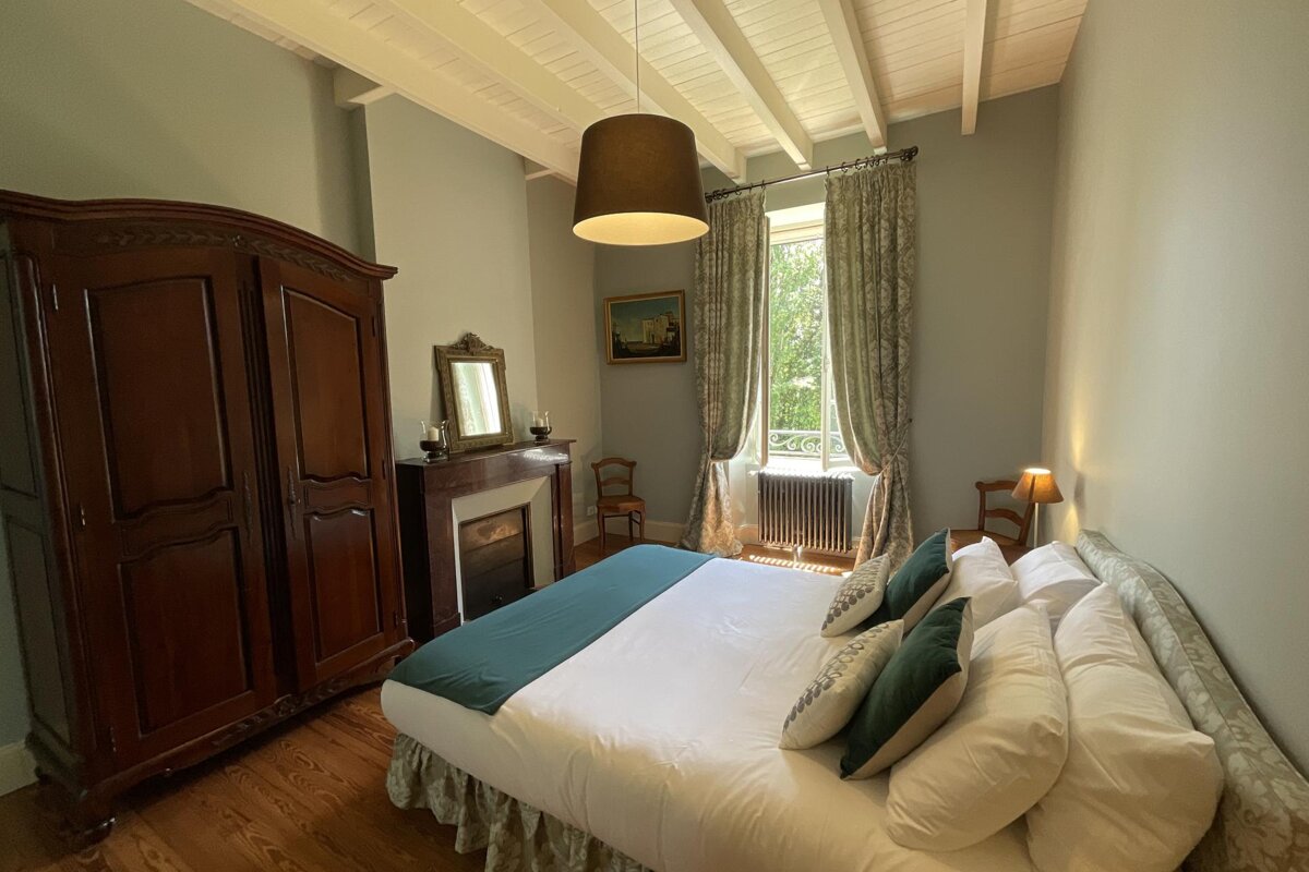 A tranquil bedroom featuring a large bed, antique wooden wardrobe, fireplace, and a bright window. Exposed ceiling beams and patterned curtains create a cozy feel.
