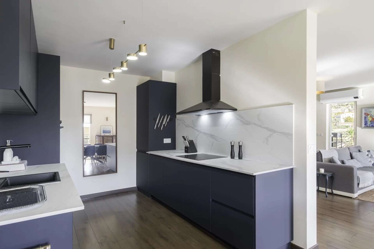 A modern navy blue and white kitchen with dark wood floors, black range hood, and integrated appliances, opening to a living room with a grey sofa.