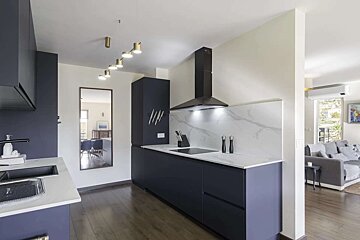 A modern navy blue and white kitchen with dark wood floors, black range hood, and integrated appliances, opening to a living room with a grey sofa.