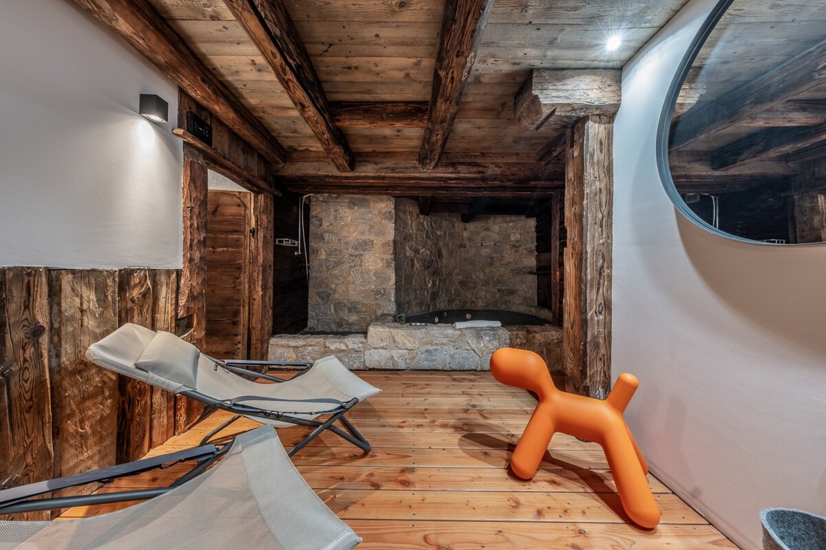 A cozy, rustic room features wooden beams, a stone hearth, and two recliners. A modern orange dog-shaped stool and round mirror add whimsy.