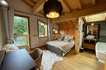 Cozy wood chalet bedroom featuring a bed, open bathroom with tub, study desk, and large windows overlooking lush trees.