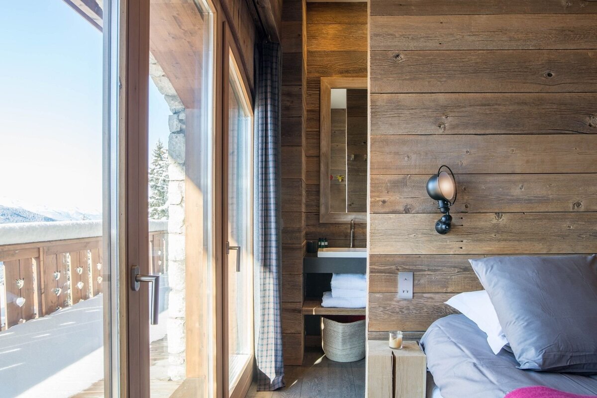 Cozy room with a large window offering snowy mountain views, rustic wood-paneled walls, a bed, and a glimpse of a bathroom.