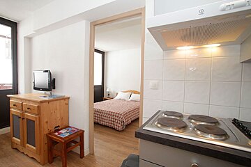 A compact studio apartment features a kitchen with a stove and range hood, a living area with a TV on a wooden cabinet, and a bedroom with a plaid bedspread visible through a doorway.